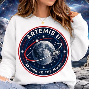 Może przedstawiać: Biała bluza z okrągłym dekoltem i okrągłą grafiką. Grafika zawiera napis "ARTEMIS II RETURN TO THE MOON", niebieski i biały księżyc, satelitę oraz czerwono-biały pierścień. Bluza jest w stylu casual.