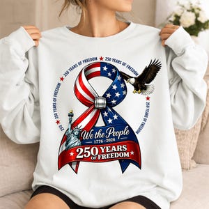 Puede incluir: Sudadera blanca con un diseño patriótico que celebra 250 años de libertad. El diseño incluye una cinta en rojo, blanco y azul, un águila, la Estatua de la Libertad y el texto "We the People 1776-2026".