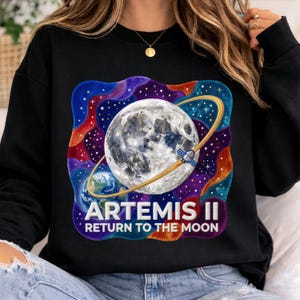 Puede incluir: Sudadera negra con un gráfico colorido de la luna, la Tierra y un satélite. El diseño incluye un anillo dorado alrededor de la luna y el texto "ARTEMIS II RETURN TO THE MOON" en blanco.