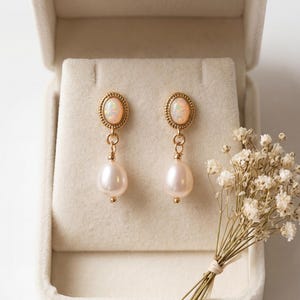 May include: Gold-coloured drop earrings featuring opal-style tops and pear-shaped pearls. The earrings are presented in an open, cream-coloured jewellery box. A small bouquet of dried flowers is to the right.