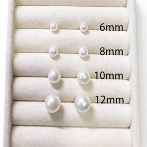 May include: A selection of pearl stud earrings in sizes 6mm, 8mm, 10mm, and 12mm. The earrings are displayed in pairs on a white, velvet-lined tray. The sizes are labelled in black text.