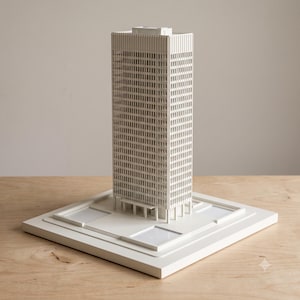 May include: A white architectural scale model of a rectangular skyscraper featuring a grid of vertical windows and a flat roof. The building stands on a square base with recessed rectangular sections at the corners and a central entrance canopy.