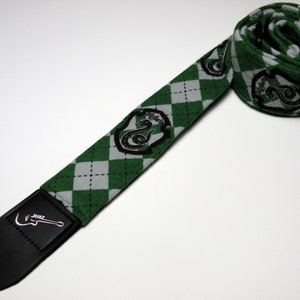 May include: Green and gray diamond-patterned guitar strap with the Slytherin crest. The strap has a black leather end with the "Jamz" logo. The strap is rolled up, showing the pattern and crest.