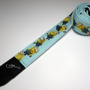 May include: A blue guitar strap with a repeating pattern of yellow Minions. The strap has a black leather end with a guitar logo.