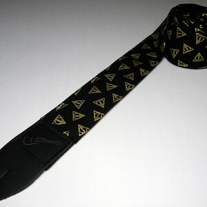 May include: Black guitar strap with a gold pattern of the Deathly Hallows symbol from the Harry Potter series.