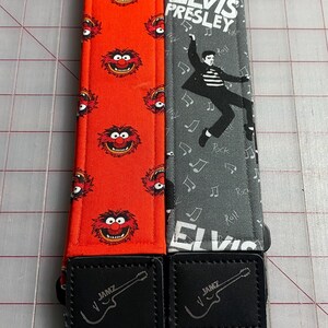 May include: Two guitar straps. One is red with a repeating cartoon face pattern. The other is gray with an Elvis Presley graphic and musical notes. Both have black leather ends with a guitar logo and the word "JAMZ".