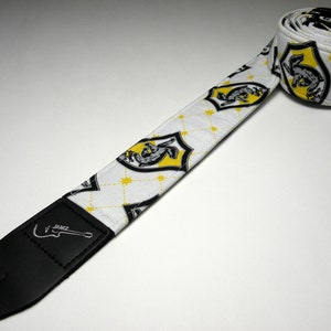 May include: White guitar strap with a black and yellow repeating pattern of a crest with a mythical creature. The strap has a black leather end with the word "JAMZ" printed on it.