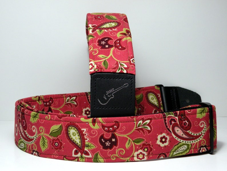 Pu&ograve; includere: Una tracolla per chitarra rossa con motivo paisley e dettagli verdi e bianchi. La tracolla ha un'estremit&agrave; in pelle nera con la scritta "JAMZ" e una silhouette di chitarra. Per chitarre elettriche.