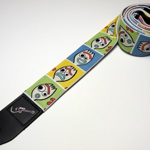 May include: A guitar strap with a colorful pattern of repeating cartoon faces. The strap is black leather with a guitar logo and the text "J.A.M.Z."
