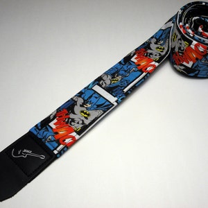 May include: A black guitar strap with a white background and a repeated pattern of Batman comic book images. The strap has a black leather end with the word "ZAKK" printed on it.