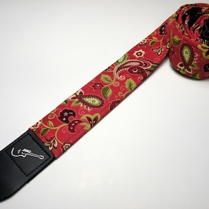 Pu&ograve; includere: Un cinturino per chitarra rosso paisley con un'estremit&agrave; in pelle nera. Il cinturino presenta un motivo ripetuto di disegni paisley verdi, bordeaux e bianchi su sfondo rosso. L'estremit&agrave; in pelle nera ha la scritta "JAMZ" e una grafica di chitarra.