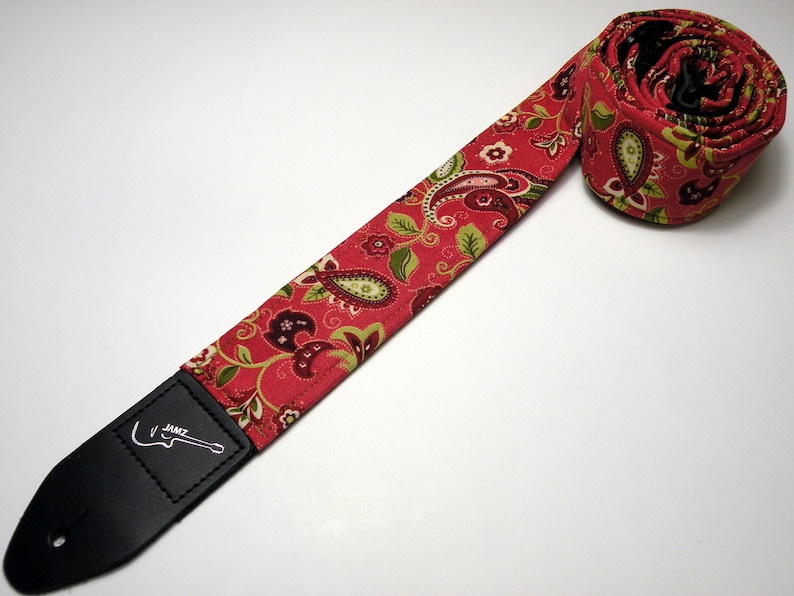Pu&ograve; includere: Un cinturino per chitarra rosso paisley con un'estremit&agrave; in pelle nera. Il cinturino presenta un motivo ripetuto di disegni paisley verdi, bordeaux e bianchi su sfondo rosso. L'estremit&agrave; in pelle nera ha la scritta "JAMZ" e una grafica di chitarra.