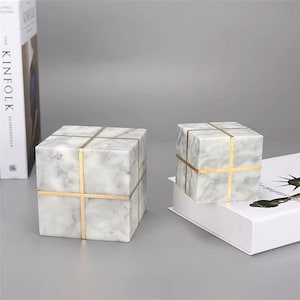 May include: Two marble cubes with gold-colored cross accents. The cubes have a white and gray veined pattern. One cube is on a white book, and the other is on a gray surface.