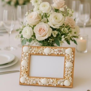 Puede incluir: Un marco de fotos de madera decorado con conchas marinas y estrellas de mar. El marco rodea un espacio en blanco, y está sobre un mantel blanco. Un arreglo floral de rosas blancas y rosas está en el fondo.