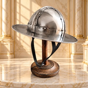 Può includere: Un elmo a cappello di ferro in acciaio lucido con tesa larga e calotta arrotondata rinforzata da fasce metalliche e rivetti. L'elmo include un sottogola in pelle nera ed è esposto su un supporto in legno con base circolare.
