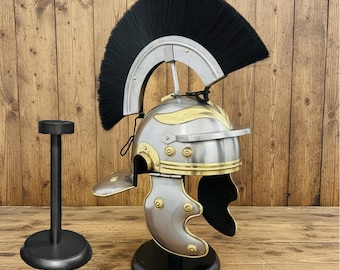 May include: A silver and gold Roman helmet with a black plume, displayed on a black stand. The helmet features brass accents and is set against a wooden background. A separate black stand is also visible.