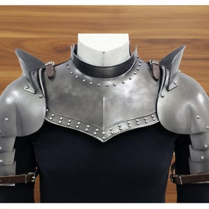 Könnte beinhalten: Ein silberfarbener mittelalterlicher Brustpanzer und Schulterpanzer. Die Rüstung besteht aus Metall und verfügt über Nieten und Lederriemen. Die Schulterpanzer haben ein mehrschichtiges Design und sind am Brustpanzer befestigt. Die Rüstung wird auf einem schwarzen Oberteil präsentiert.