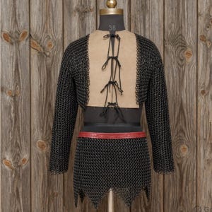 May include: A full suit of chainmail armor displayed on a mannequin. The armor is black with a tan backing and a red belt. The mannequin is set against a wooden background.
