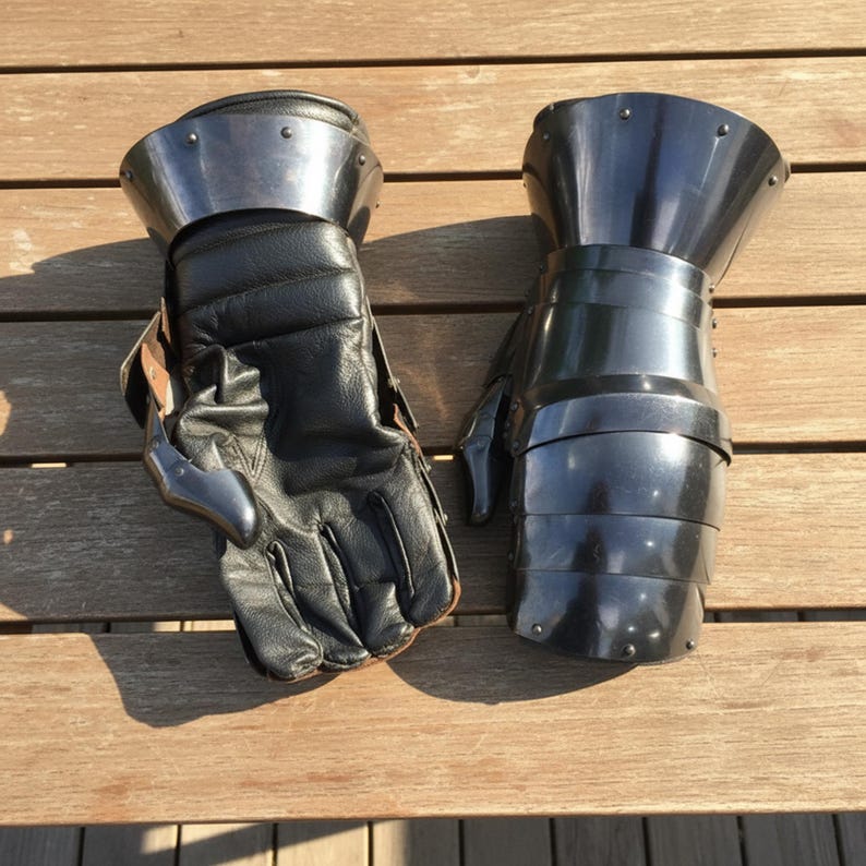 May include: A pair of medieval gauntlets, one with a black leather glove and steel accents, the other fully steel. The gauntlets are designed for protection and are placed on a wooden surface.