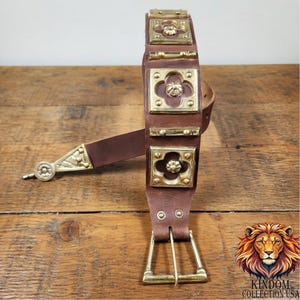 May include: A brown leather belt with gold-coloured metal embellishments. The belt has square and rectangular decorative plates, a buckle, and a decorative end piece. The belt is displayed on a wooden surface.