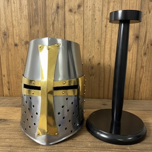 Peut inclure: Un casque médiéval argenté avec une croix et des garnitures dorées, à côté d'un support de casque noir. Le casque comporte de petits trous de ventilation et un fond pointu. Le support a une base ronde et un poteau cylindrique.