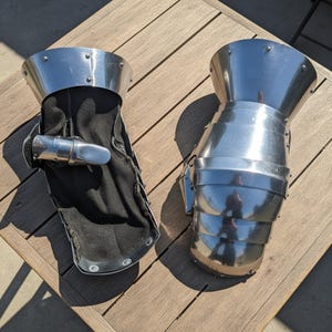 May include: Two pieces of medieval-style gauntlets. The gauntlets are made of polished metal and feature articulated plates for flexibility. One gauntlet has a black interior lining. The gauntlets are designed to protect the hands and wrists.