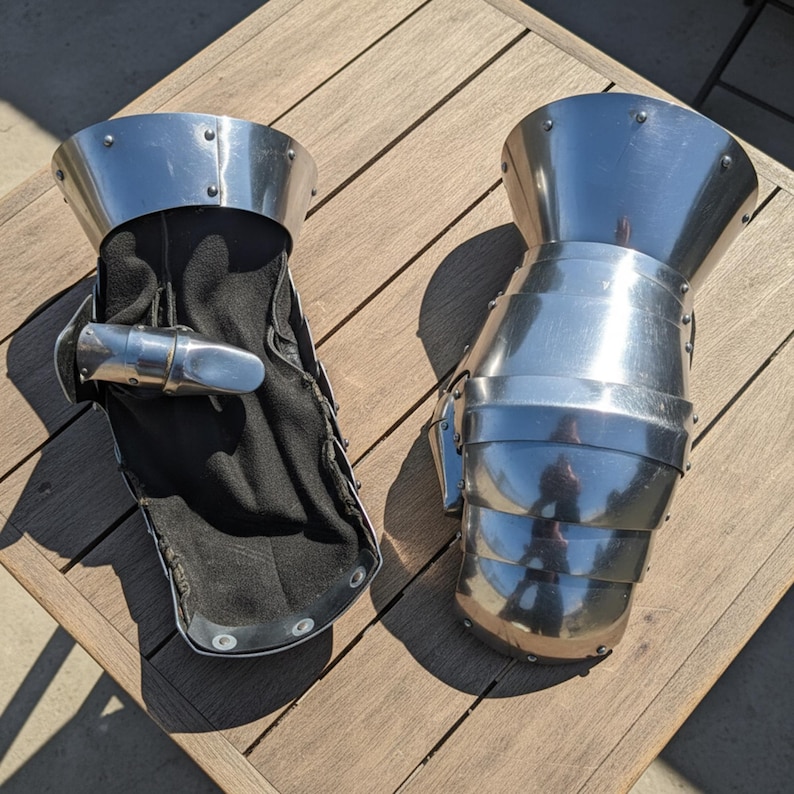 May include: Two pieces of medieval-style gauntlets. The gauntlets are made of polished metal and feature articulated plates for flexibility. One gauntlet has a black interior lining. The gauntlets are designed to protect the hands and wrists.
