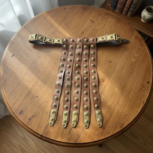 May include: A brown leather Roman arm guard with brass studs and accents. The arm guard is displayed on a wooden table. The design features multiple leather straps with brass studs and rectangular brass plates.