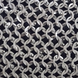 May include: Close-up of chainmail armour. The silver-coloured metal rings are interlocked in a dense, overlapping pattern. The background is a dark, textured fabric, contrasting with the metallic sheen of the chainmail.