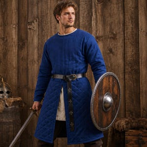 May include: A long-sleeved blue quilted tunic featuring a diamond-stitched pattern, a round neckline, and a front slit. The garment is cinched at the waist with a dark brown leather belt featuring a metal buckle and decorative metal studs.