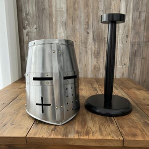 Puede incluir: Un casco de cruzado medieval plateado con un recorte de cruz y detalles de remaches, junto a un soporte de casco negro. El casco tiene ranuras para los ojos y orificios de ventilación. El soporte es redondo en la base y en la parte superior.