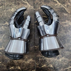 May include: Pair of steel gauntlets with articulated fingers and leather palms. The gauntlets have a polished metal finish, riveted construction, and brown leather accents. The design is reminiscent of medieval armor.