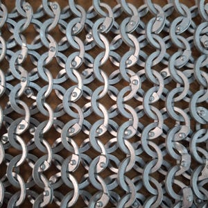 May include: Close-up of a section of chainmail armour. The interlocking metal rings are silver-coloured and arranged in a dense pattern. Each ring is riveted, creating a durable and flexible mesh. The background is a warm, neutral tone.