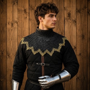May include: A black quilted gambeson featuring a high collar and a black chainmail aventail with a scalloped gold-toned metal ring border. The garment includes a central leather strap with a metal buckle and polished steel gauntlets on the arms.