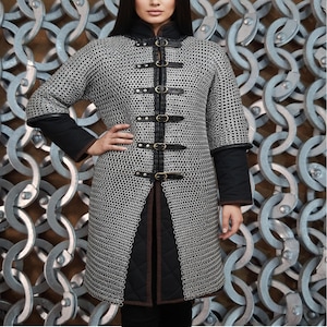 May include: A silver chainmail coat with black leather accents and buckles. The coat has short sleeves and a high collar. The front of the coat is open, revealing a black quilted underlayer. The coat is designed for historical reenactment or costume use.