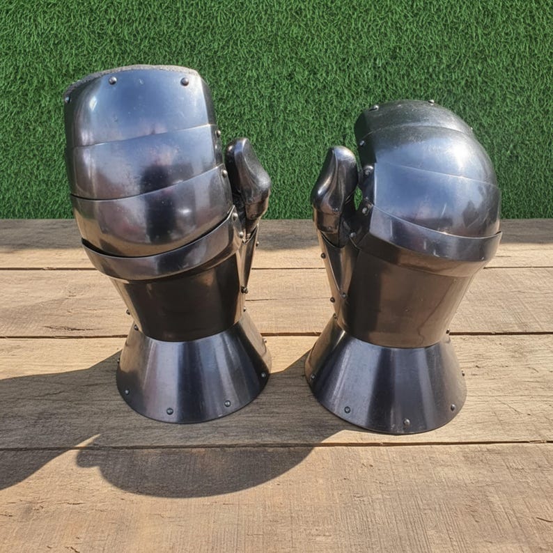 May include: Pair of polished steel gauntlets, likely for historical reenactment or display. The gauntlets are dark gray with articulated fingers and a flared cuff. They are set against a wooden surface with a green background.