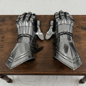 May include: A pair of polished steel gauntlets, featuring articulated fingers and wrist protection. The medieval-style armor gloves are designed with overlapping plates and leather straps. The gauntlets are displayed on a wooden surface.