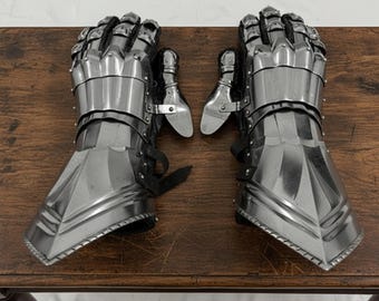 May include: A pair of polished steel gauntlets, featuring articulated fingers and wrist protection. The medieval-style armor gloves are designed with overlapping plates and leather straps. The gauntlets are displayed on a wooden surface.