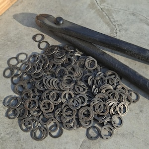 May include: A pile of small, circular metal washers with a keyhole-shaped opening, alongside a large, black metal tool with long handles. The washers are scattered on a concrete surface, and the tool is positioned diagonally across the top.