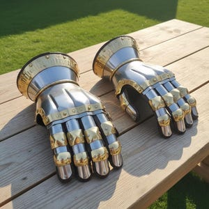 May include: A pair of steel and brass gauntlets, designed to protect the hands. The gauntlets feature articulated fingers and a flared cuff with brass accents. The metal gloves are displayed on a wooden surface, showcasing their craftsmanship.