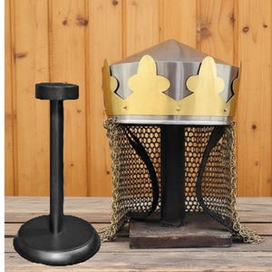 Può includere: Un elmo medievale con un design a corona dorata e cotta di maglia, esposto su un supporto nero. L'elmo ha una parte superiore argentata ed è posto su una superficie di legno. È visibile anche un supporto nero separato.