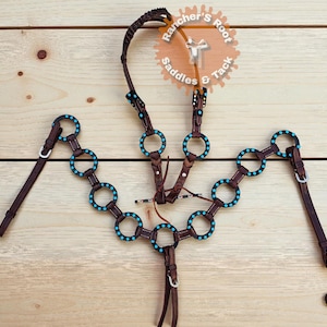 May include: A brown leather horse breast collar with turquoise accents. The collar features connected rings with turquoise studs, and adjustable straps. The image also shows the text "Rancher's Root Saddles & Tack".