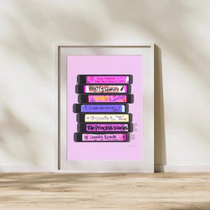 May include: A framed art print on a light pink background featuring a stack of seven black VHS tapes. The labels display titles including Romy and Michele's High School Reunion, Pretty Woman, Clueless, 10 Things I Hate About You, Dirty Dancing, The Princess Diaries, and Legally Blonde.
