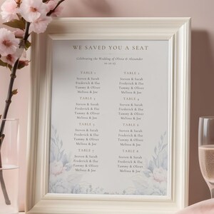 Könnte beinhalten: Ein gerahmter Hochzeits-Sitzplan mit dem Text "WE SAVED YOU A SEAT". Der Plan listet Namen unter Tischnummern mit floralen Akzenten auf. Ein Glas Champagner und rosa Blumen sind im Hintergrund.