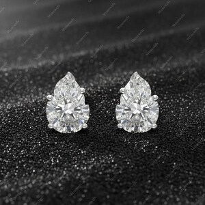 May include: A pair of pear-cut diamond stud earrings. The diamonds are set in a silver or white gold setting, sparkling against a dark, textured background. A classic jewellery item.