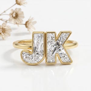 May include: A gold ring featuring two large, clear, faceted letter-shaped stones forming the initials JK. The letters are set in a polished gold band, with the text Stonewels engraved on the inner side of the band, showcasing a clear, reflective design.