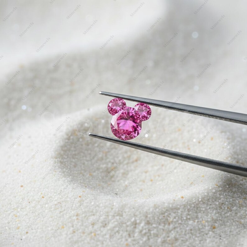 May include: A pink gemstone in the shape of a mouse head, held by tweezers against a white, textured backdrop. The gemstone features a large, round centre and two smaller, round gems for ears. The tweezers are silver.