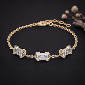 Könnte beinhalten: Ein goldenes Kettenarmband mit drei klaren, rechteckig geschliffenen Edelsteinen in knochenförmigen Metallfassungen. Das Armband besteht aus einer feinen Gliederkette und einem sicheren Karabinerverschluss mit einer polierten Oberfläche.