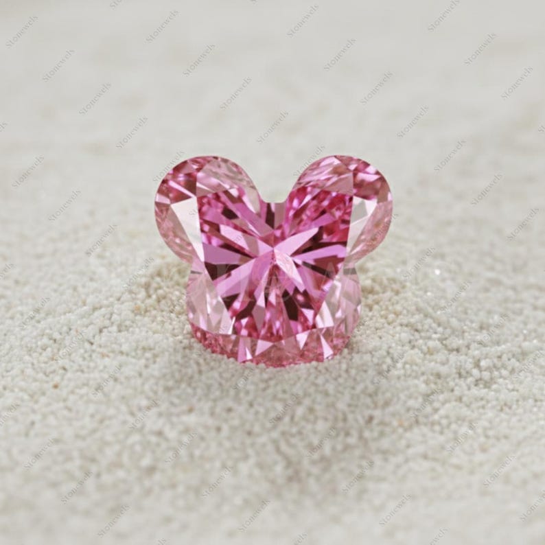 May include: A pink gemstone with a distinctive shape, possibly a bow or a stylized design. The gem is set on a textured, off-white background. The facets of the gem reflect light, creating a sparkling effect. The gem's colour is a vibrant pink.