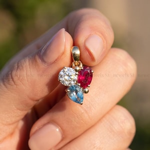 May include: A gold pendant featuring a cluster of three faceted gemstones: a round clear white stone, an oval deep red stone, and a pear-shaped light blue stone. The stones are set in a polished gold metal frame with a small bail at the top.